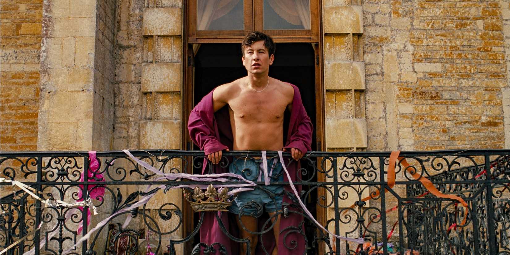 Oliver, wearing a robe, standing on a balcony and looking over the Saltburn estate in Saltburn.