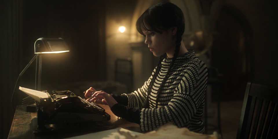 Wednesday Addams sits down to type at a typewriter while wearing a striped sweater in Wednesday season 2 episode 5.