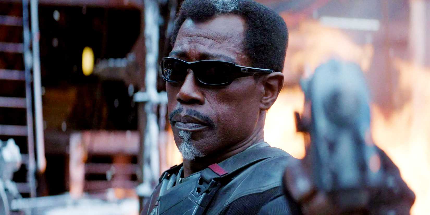 wesley snipes blade aims a gun while a fire rages behind him in deadpool wolverine