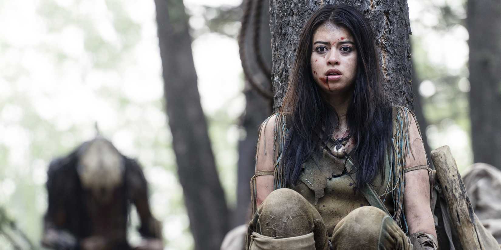 Amber Midthunder se esconde atrás de uma árvore enquanto um alienígena espreita ao fundo em uma cena de Prey.