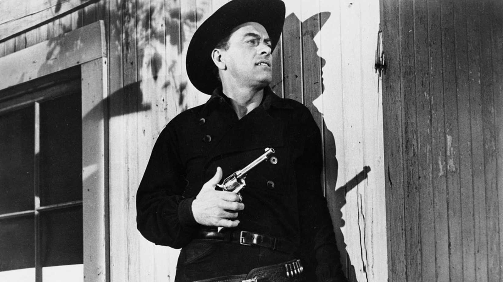 John Ireland leans against a wall with a pistol in his hand in The Gunslinger