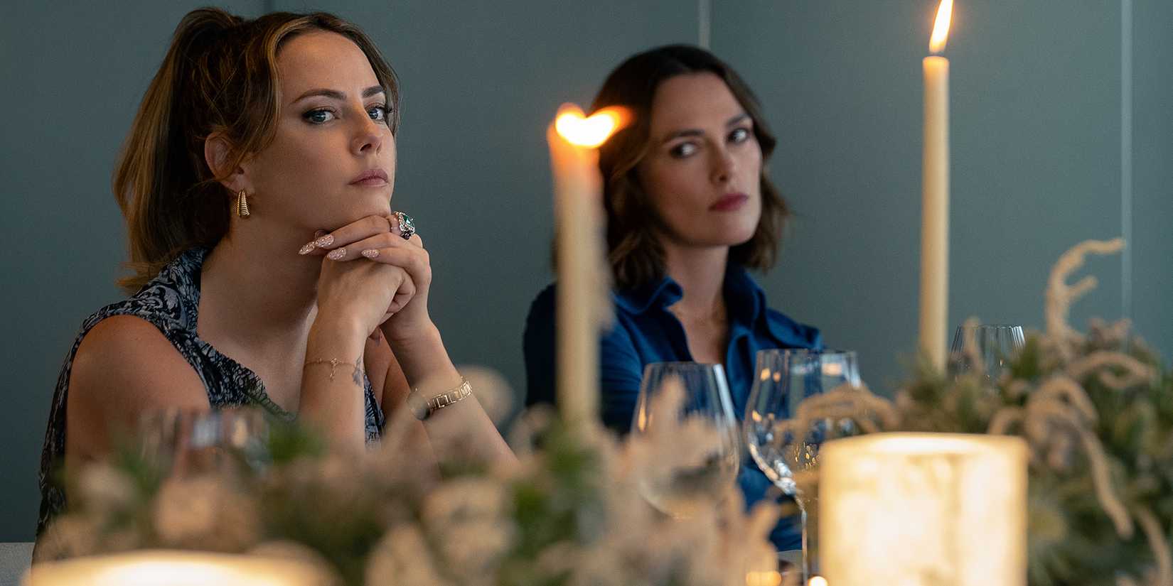 Keira Knightley's Lo looking judgmentally at Kaya Scodelario's Grace while at a table in The Woman in Cabin 10
