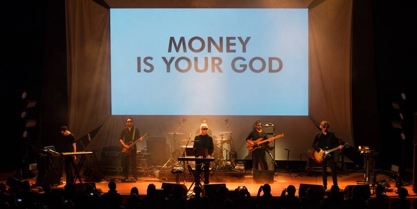 John Carpenter, Cody Carpenter, Daniel Davies, live at the Warwick Arts Centre standing in front of a large projection that says "Money is your God."