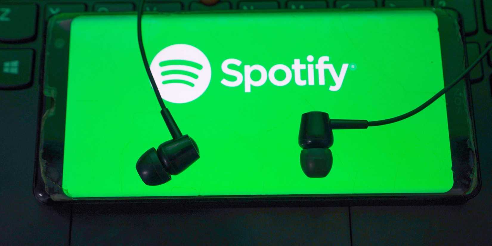 The Spotify logo is present on a green backdrop with white letters and a white circle. There are parts of a keyboard in the background and earbuds sitting on top of the logo area.