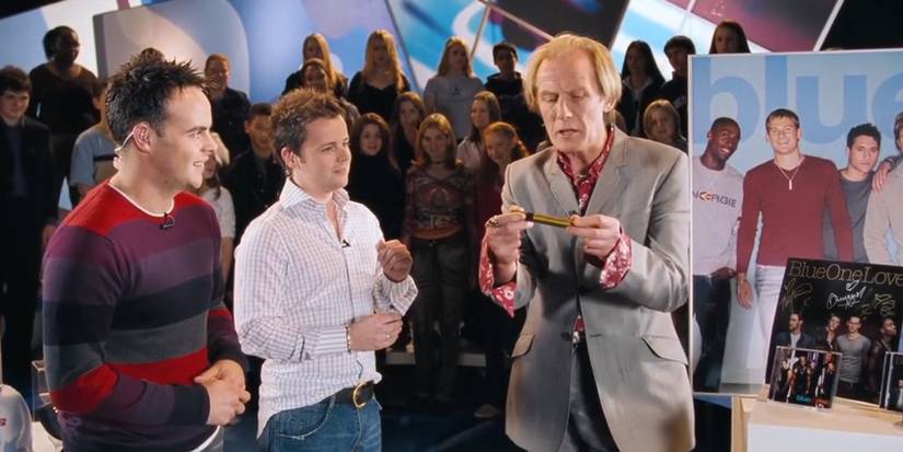 Ant and Dec looking at Billy Mack as he holds out a gold pen during an interview, with a crowd behind them, in Love Actually.