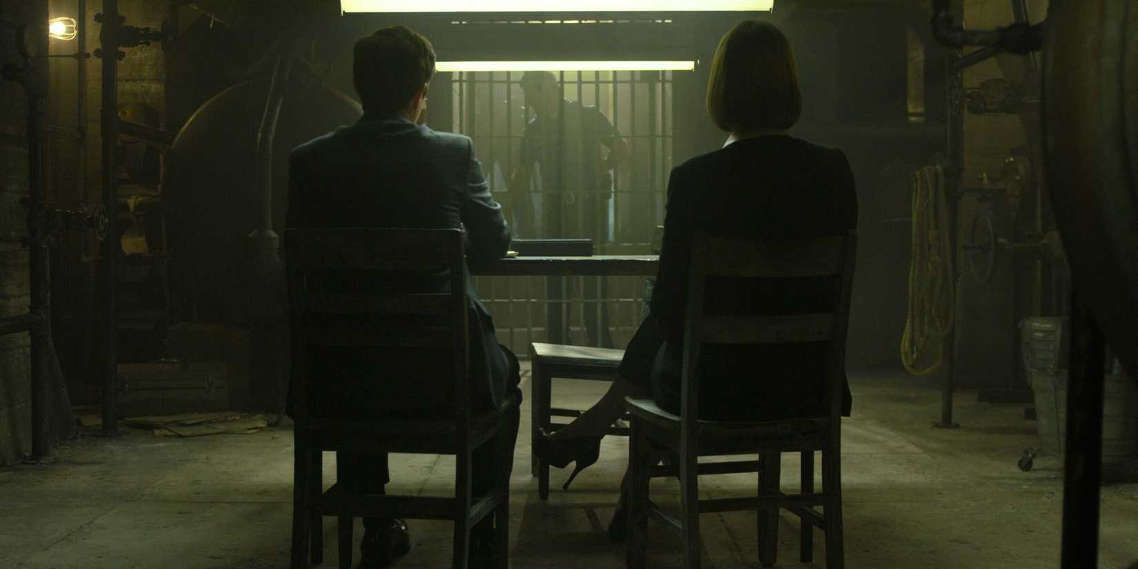 Joe Tuttle e Anna Torv sentados em uma mesa em Mindhunter, esperando por um sujeito atrás das grades da prisão.