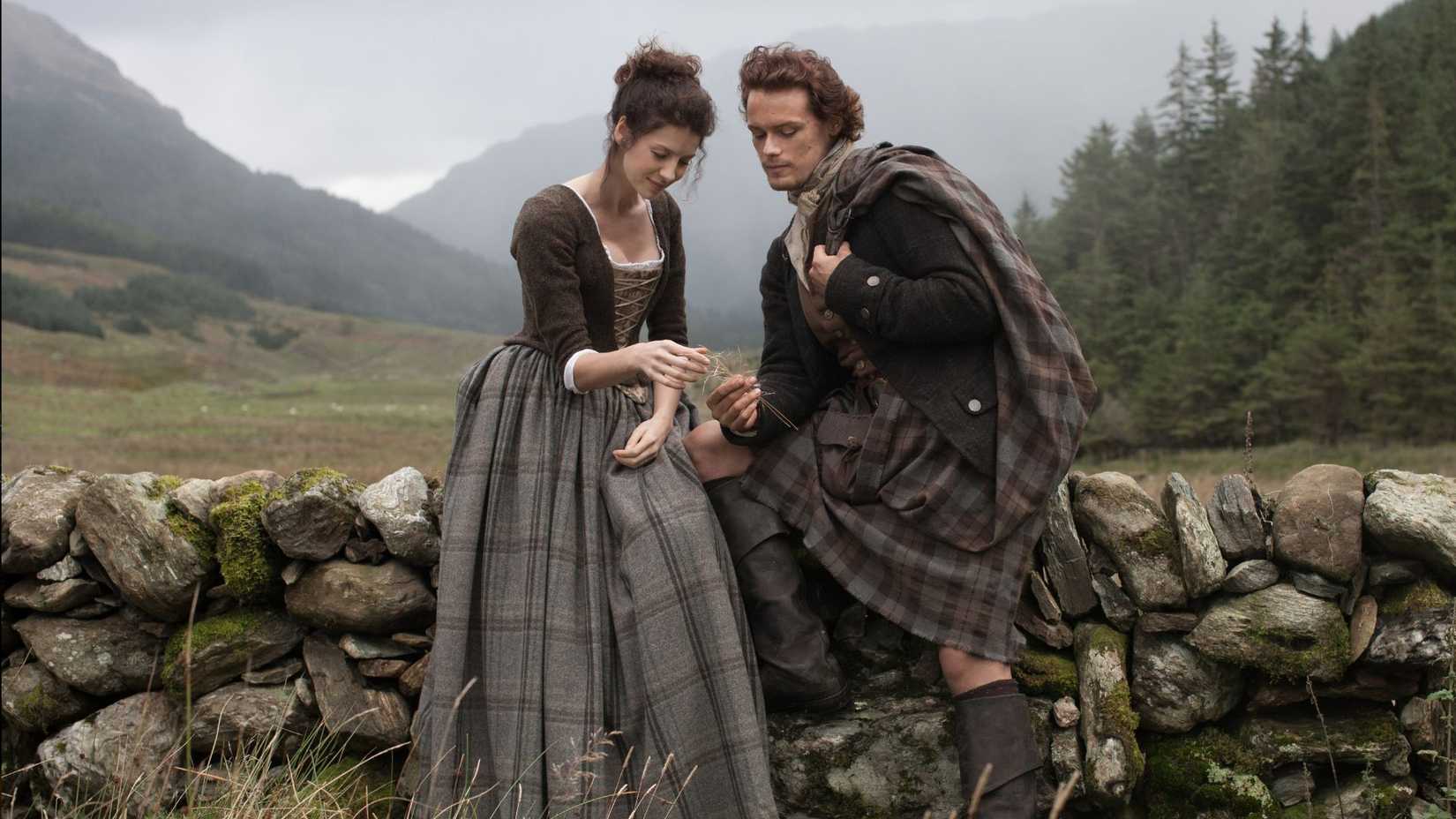 Claire and Jamie chat while sitting on a stone wall in Outlander