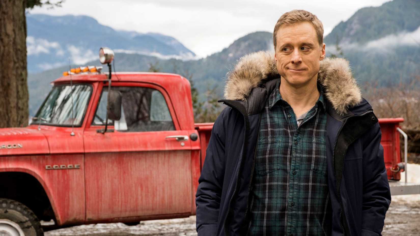 Harry dando um olhar de lado com seu caminhão e as montanhas ao fundo em Resident Alien