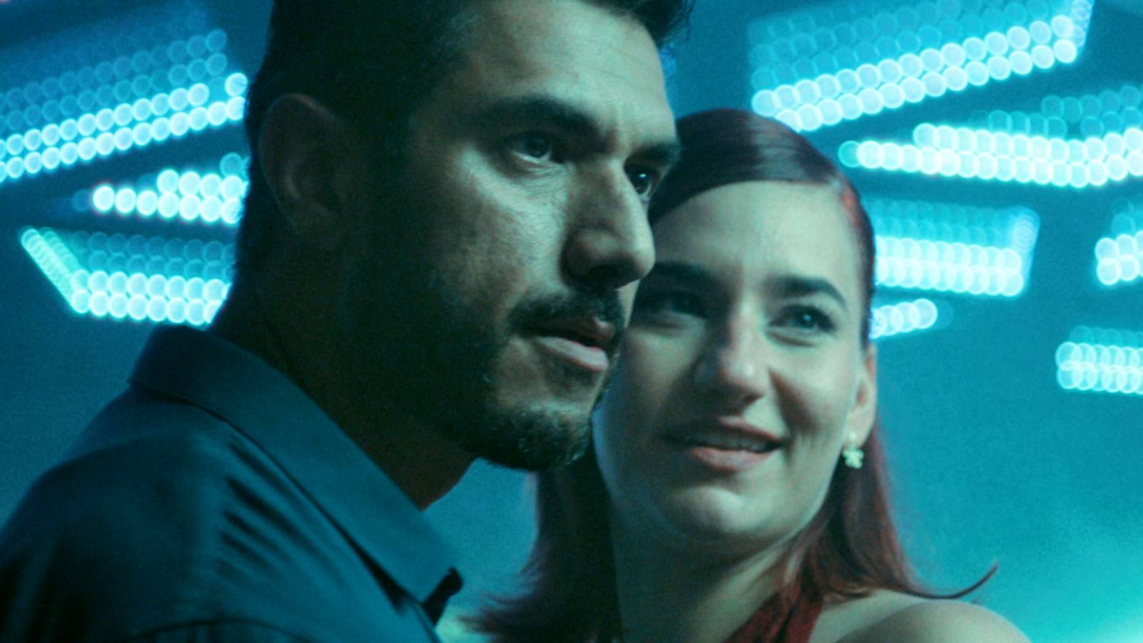 Afshin Firouzi and Maria Cordsen looking into the distance beneath a brightly lit ceiling in The Asset