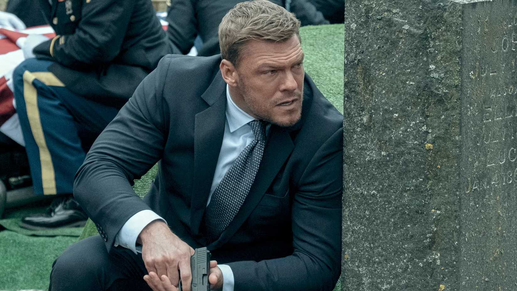 Alan Ritchson als Reacher, der sich hinter einem Grab versteckt