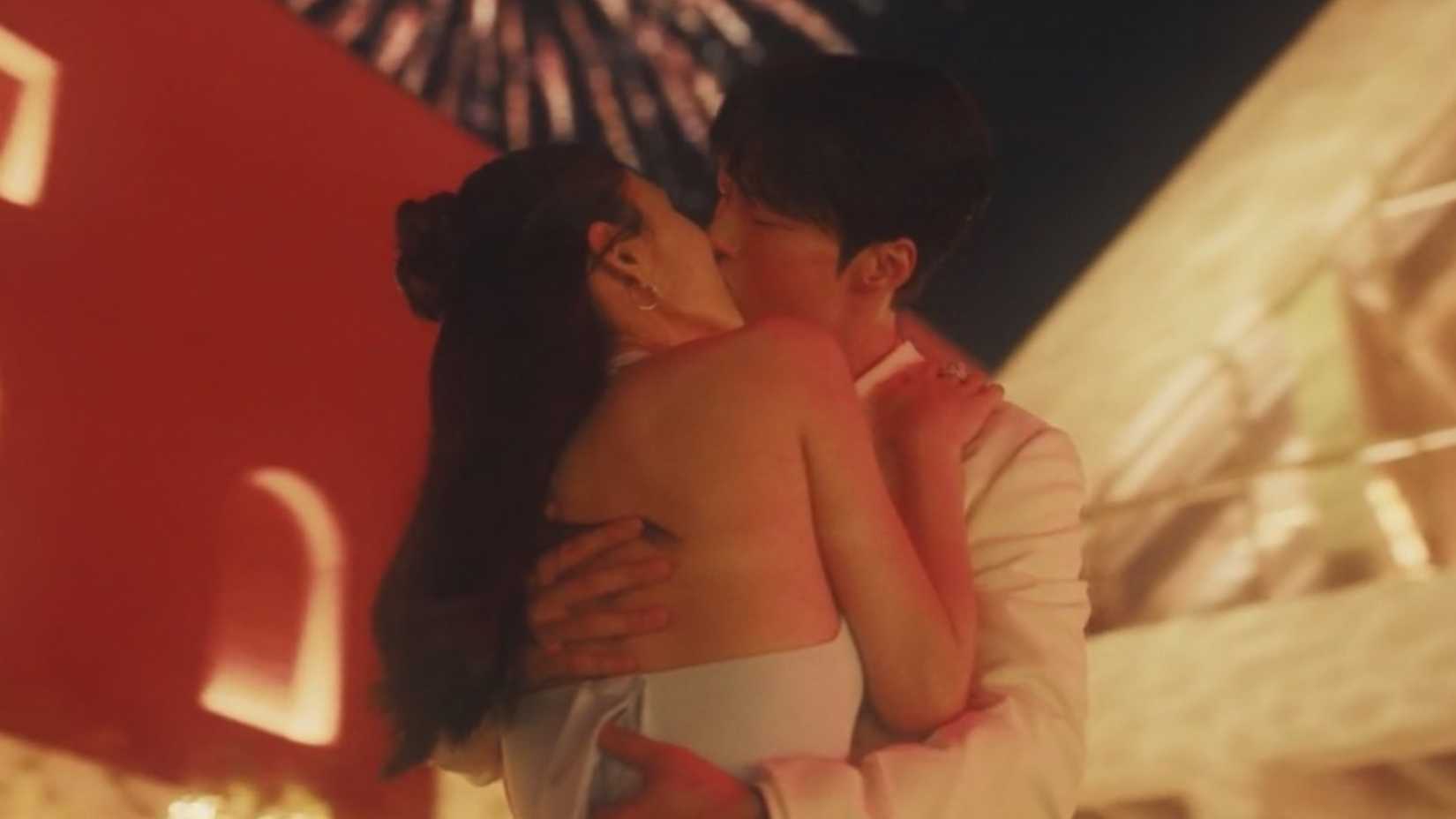 Um homem e uma mulher se beijam com fogos de artifício ao fundo.