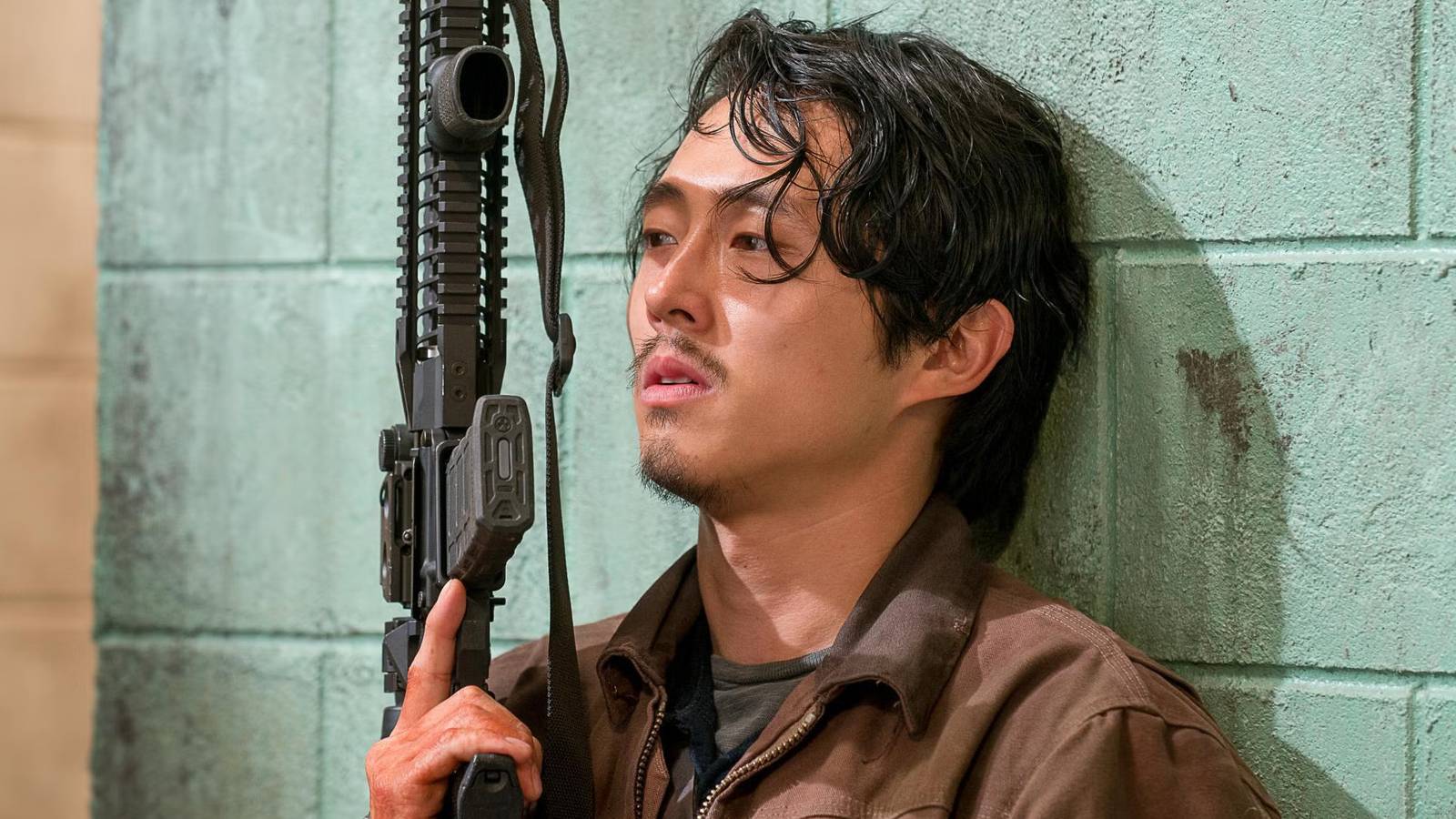 Steven Yeun as Glenn holding a gun while leaning against the wall in The Walking Dead