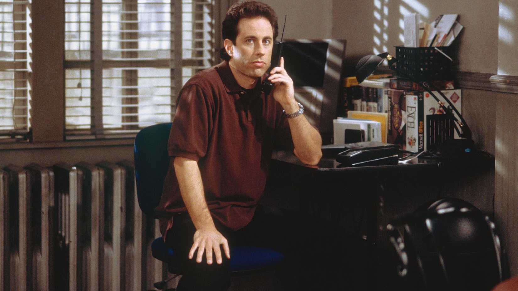 Jerry Seinfeld sentado em seu apartamento segurando um telefone