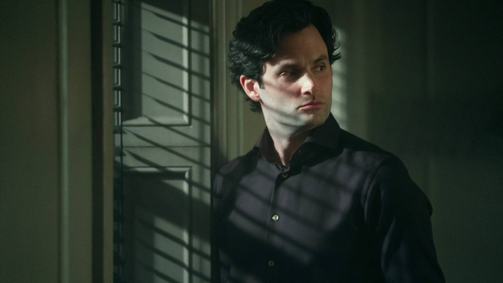 Joe Goldberg (Penn Badgley) in a shadowy room looking at something in suspicion in You season 5