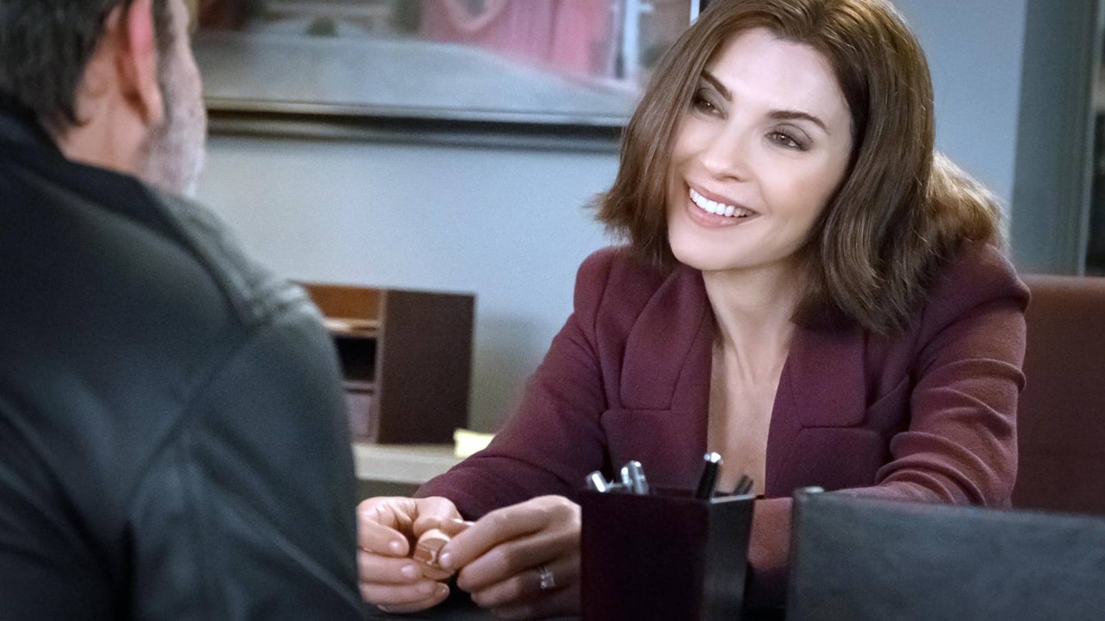 Julianna Marguiles as Alicia Florrick, sitting at a desk and smiling in The Good Wife