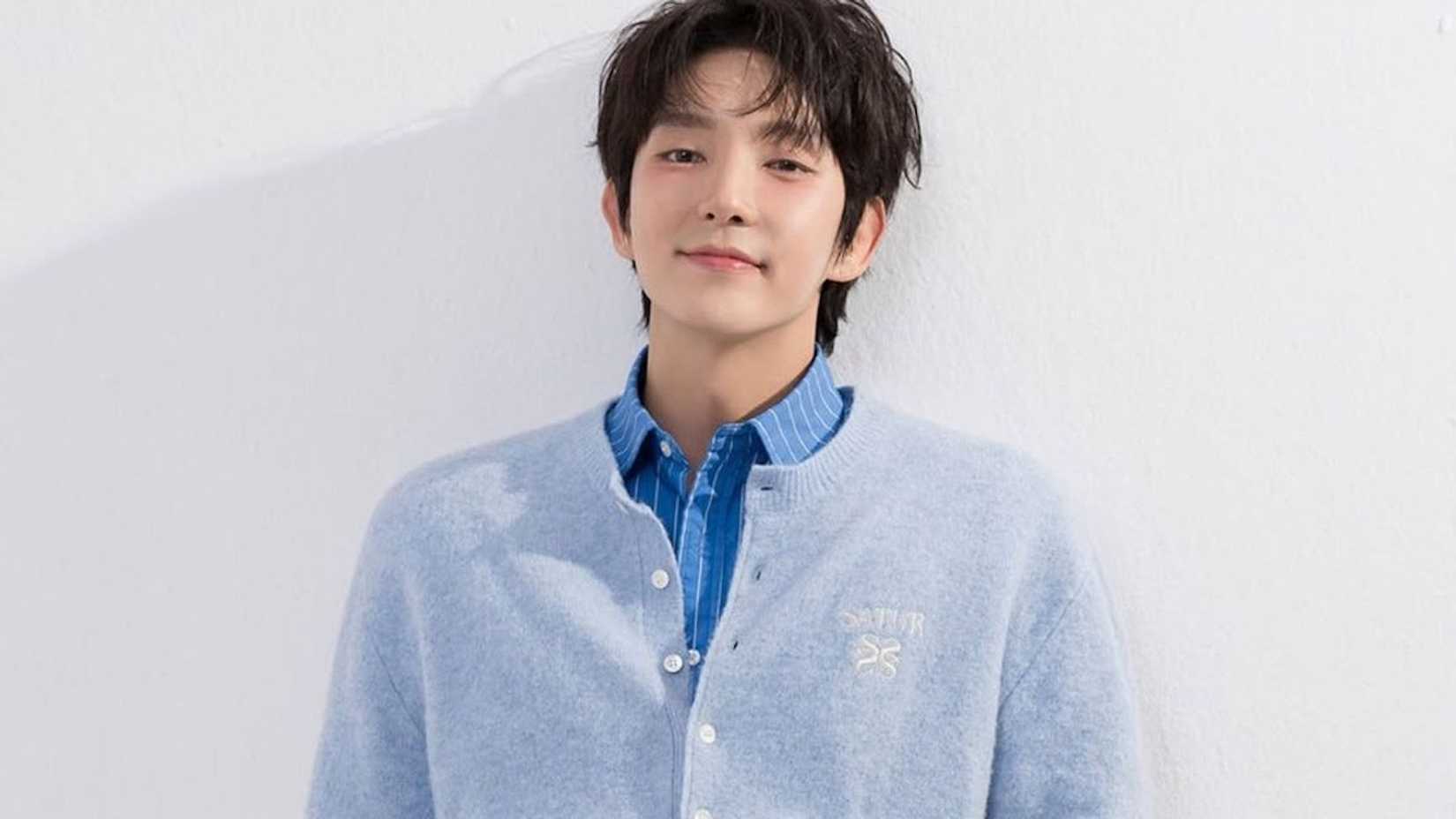 Lee Joon-gi sorrindo em uma camisa azul e suéter azul contra um fundo branco.