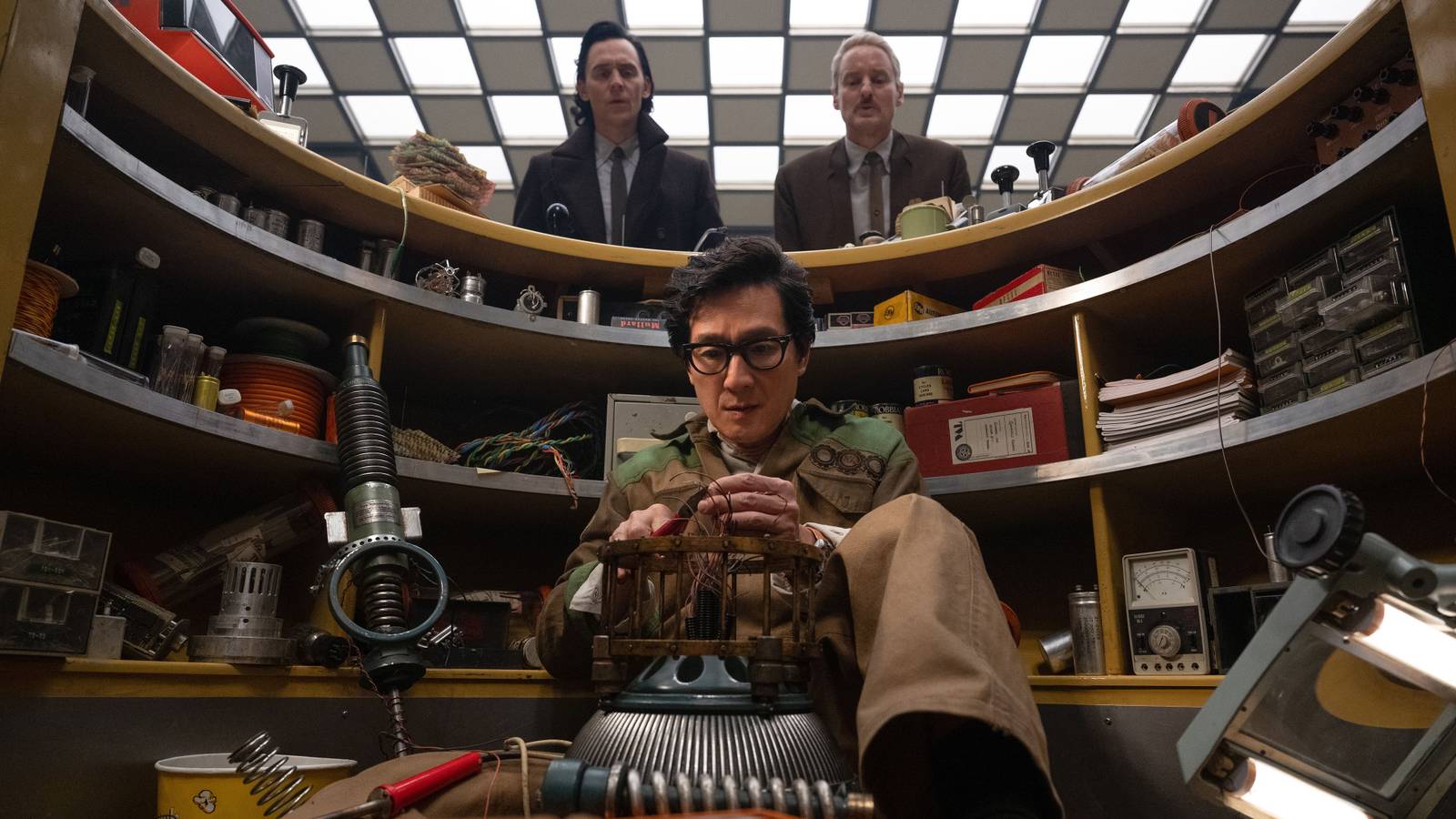 Ke Huy Quan's OB working on a device on the floor while Tom Hiddleston's Loki and Owen Wilson's Mobius peer over the desk in Loki season 2