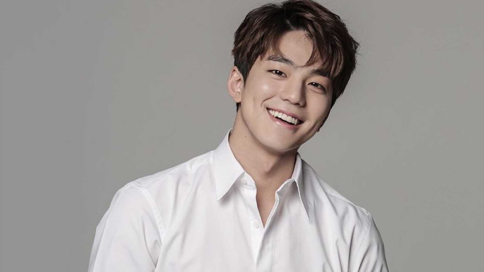 Kim Min-kyu sorrindo em uma camisa branca contra um fundo cinza.