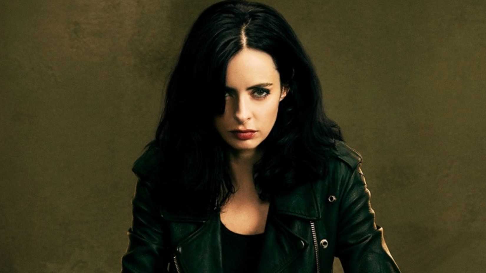 Krysten Ritter como Jessica Jones sentada em uma mesa, olhando diretamente para a câmera em um pôster da série Jessica Jones.