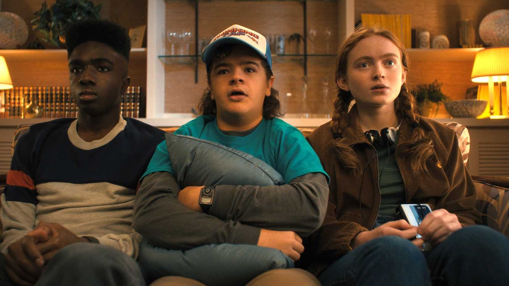 Lucas Dustin and Max sit together on a couch in Stranger Things