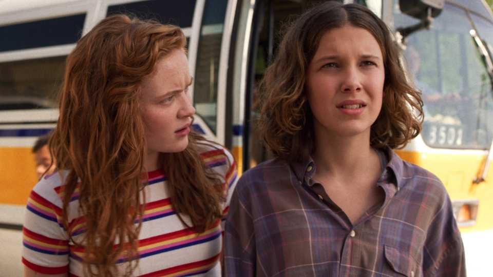 Max and Eleven standing in front of a bus in Stranger Things.