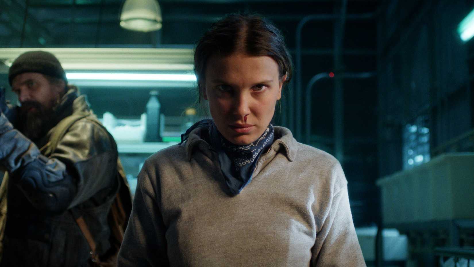 Millie Bobby Brown as Eleven in Stranger Things season 5, David Harbour as Hopper carrying a gun in the background