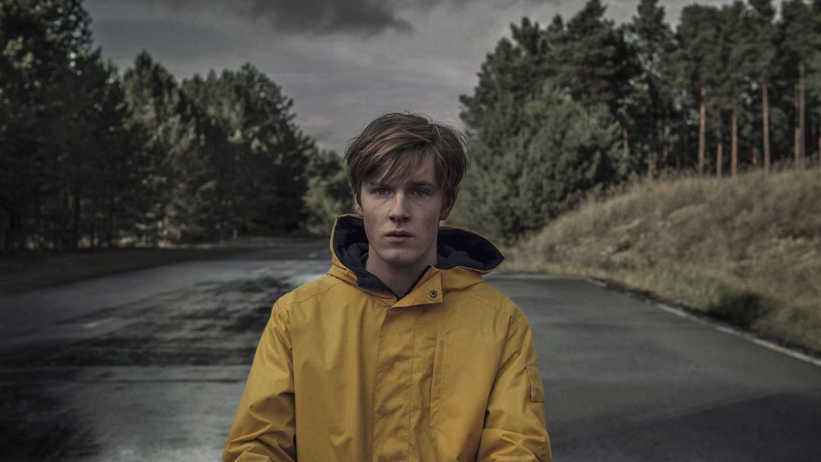 Jonas em Dark, vestindo um casaco amarelo, com a floresta atrás dele.