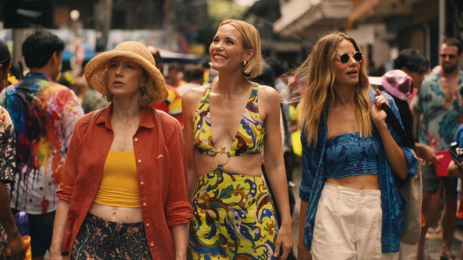 Three women in bright vacation wear walk through a crowded street in The White Lotus season 3.