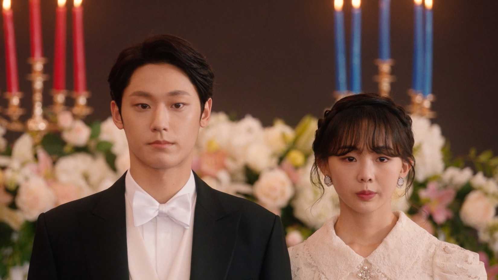 Um homem e uma mulher em trajes formais em frente a flores e velas vermelhas e azuis no K-drama Youth of May.