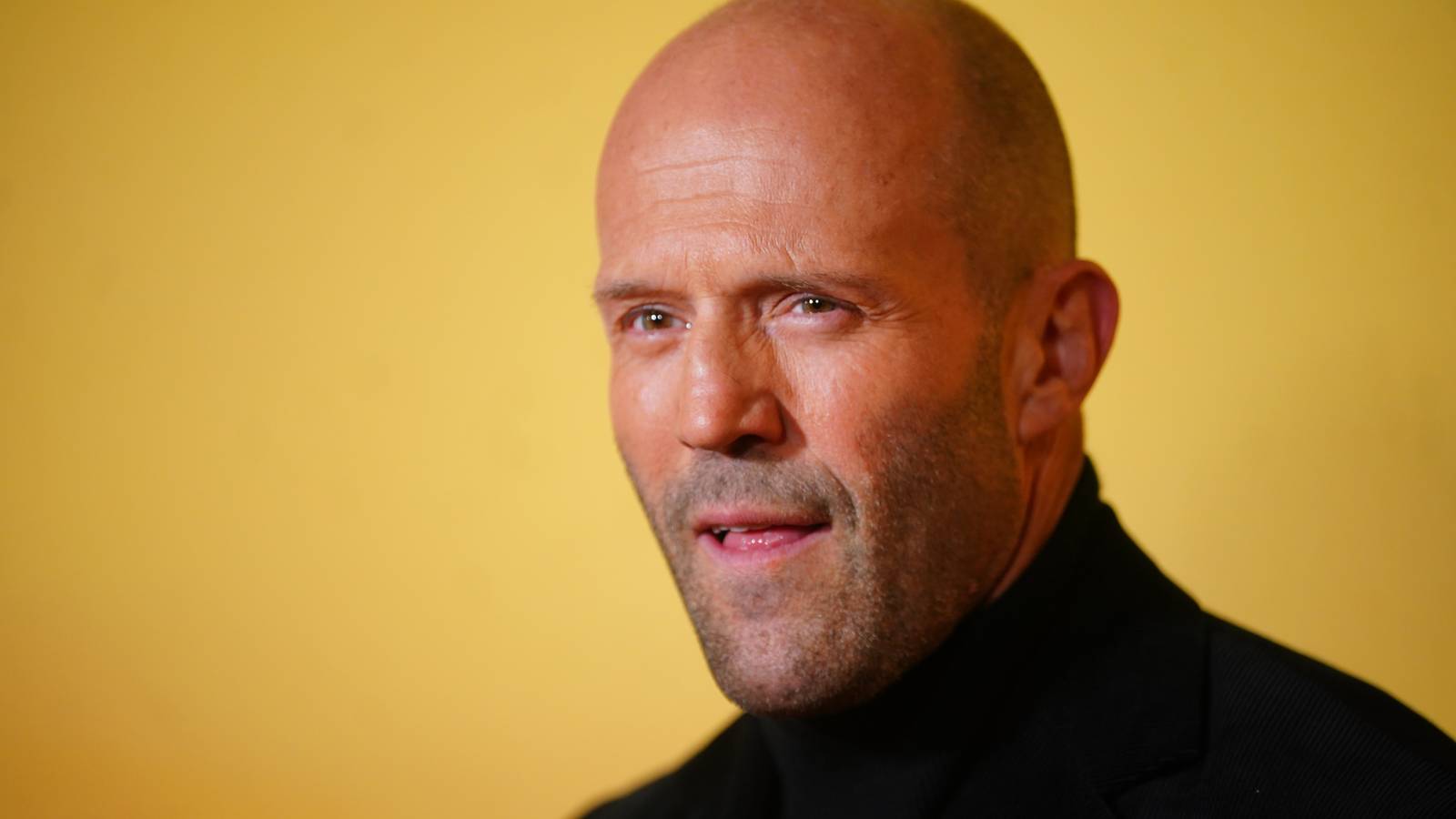 Jason Statham licks his lip while looking forward at a red carpet event.