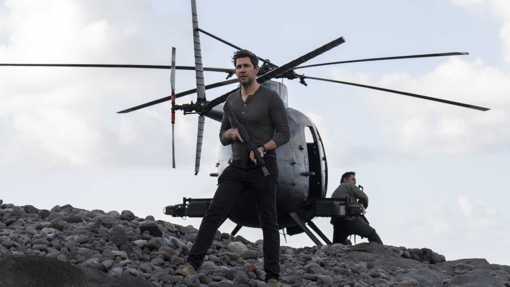 John Krasinski as Jack Ryan standing in front of a helicopter with a gun.