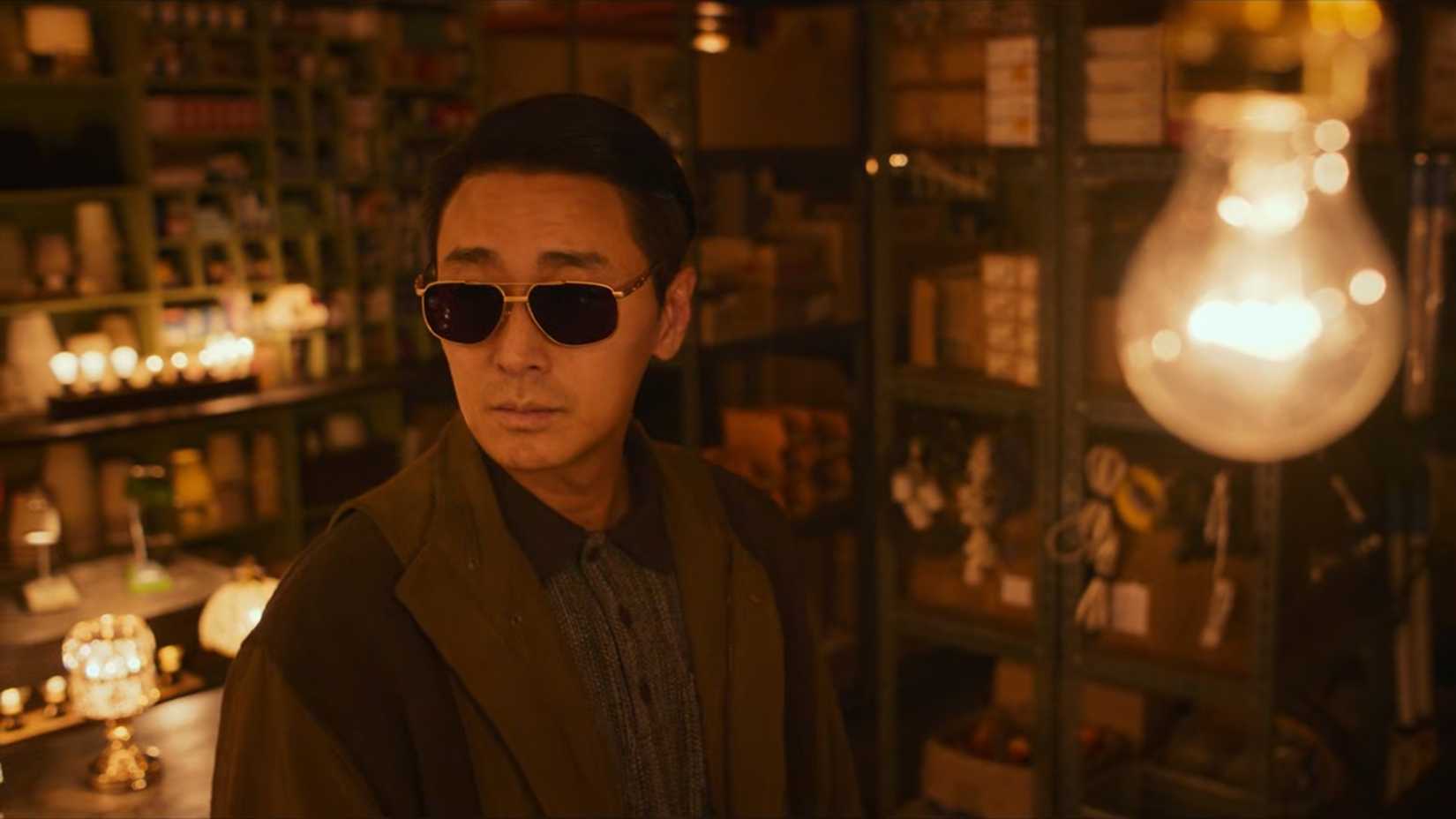 A man in sunglasses stands in a room with dim lights in the K-drama Light Shop.