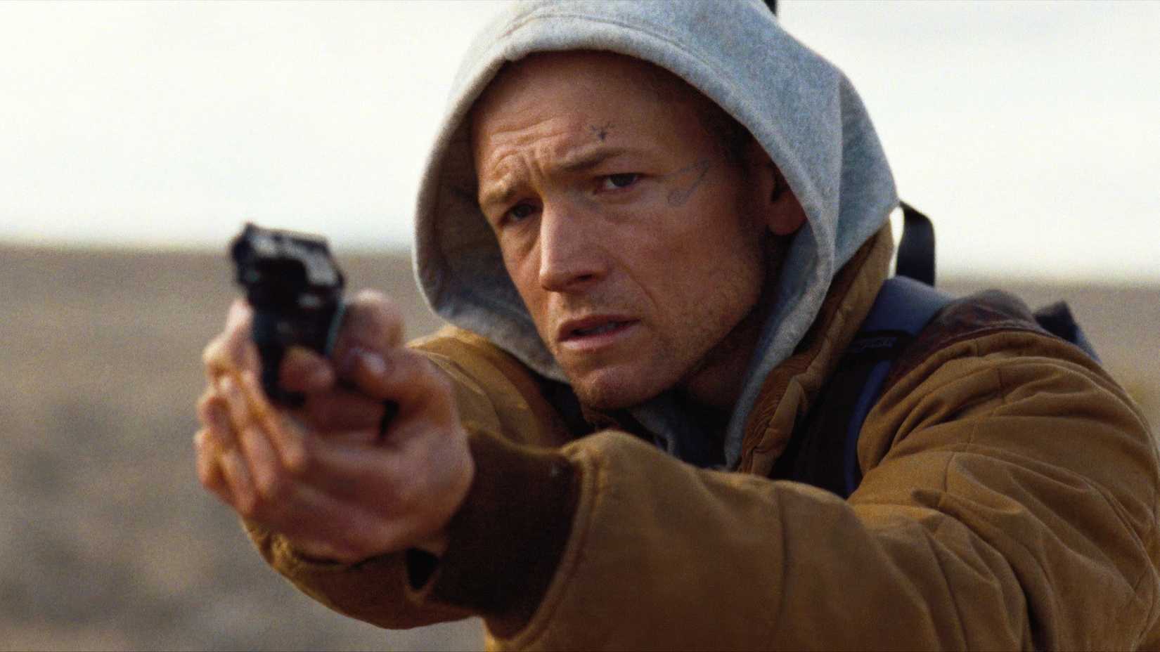 Taron Egerton pointing a gun and wearing a hoodie in She Rides Shotgun