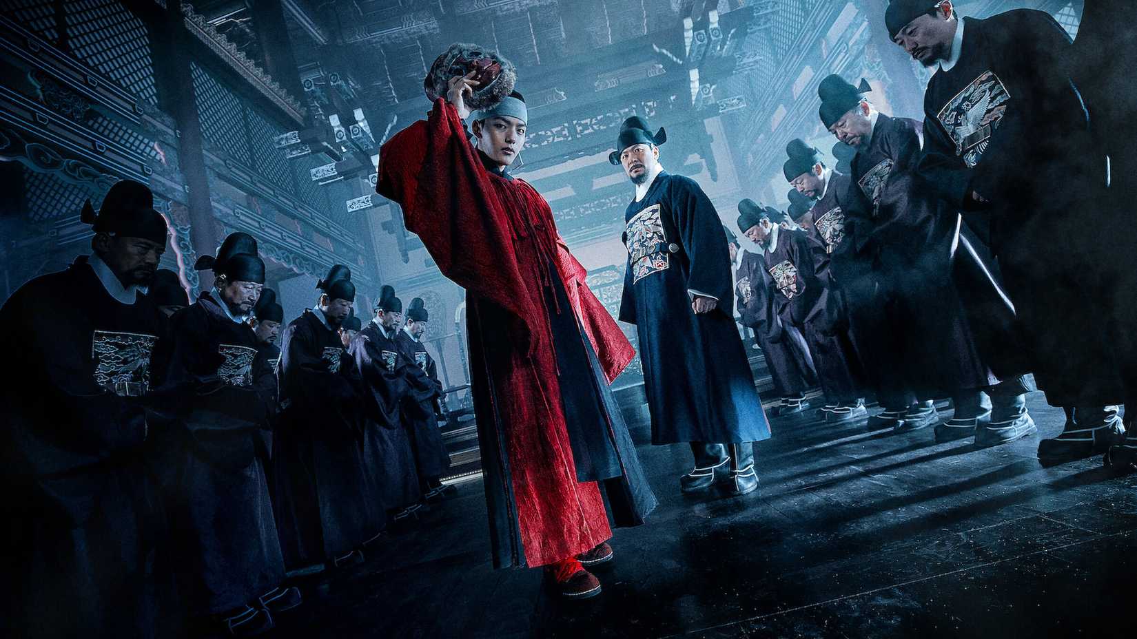 A man in red royal robes stands between two rows of bowing servants in the K-drama The Crowned Clown.
