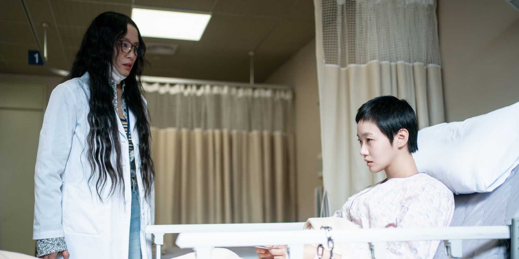 The Price Of Confession still featuring a doctor looking at a patient restrained in a hospital bed.