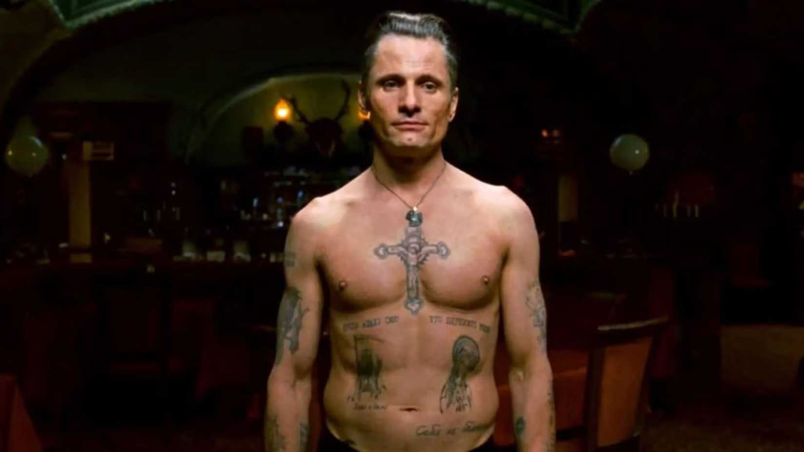 a shirtless nikolai luzhin stands inside an empty bar with his shirt off exposing his numerous gang affiliated tattoos in easter promises