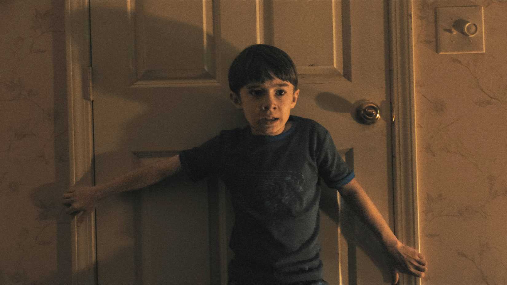 A terrified Alex holds a door shut with his back in Weapons