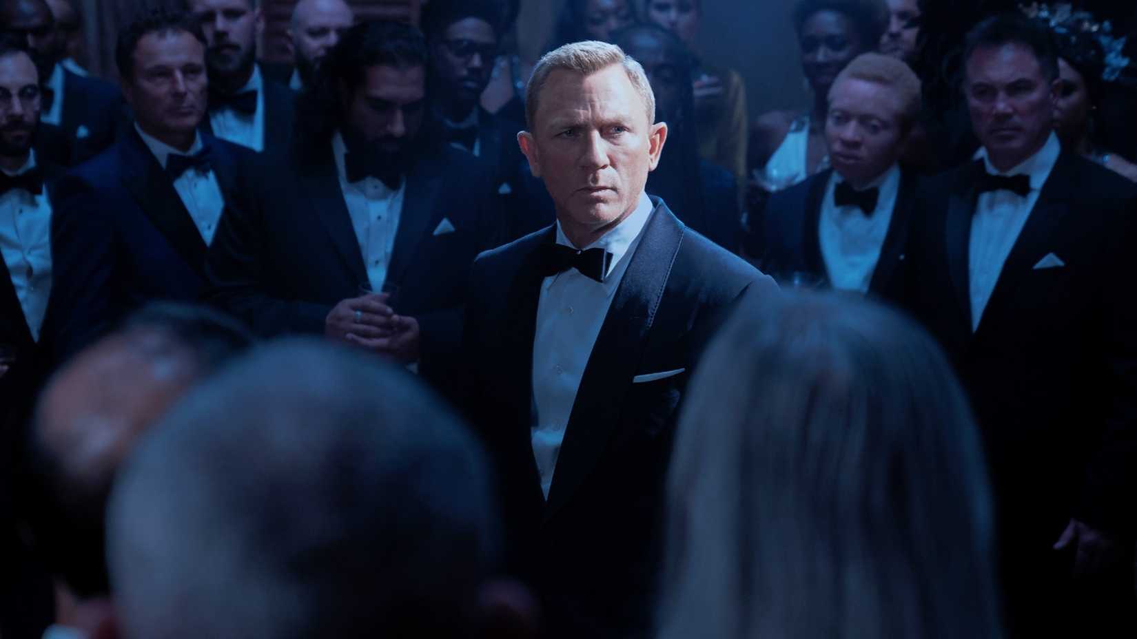 Daniel Craig as James Bond stands in a room of men in suits. 
