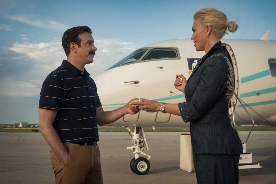 Ted Lasso and Rebecca Welton touching hands with a plane in the background