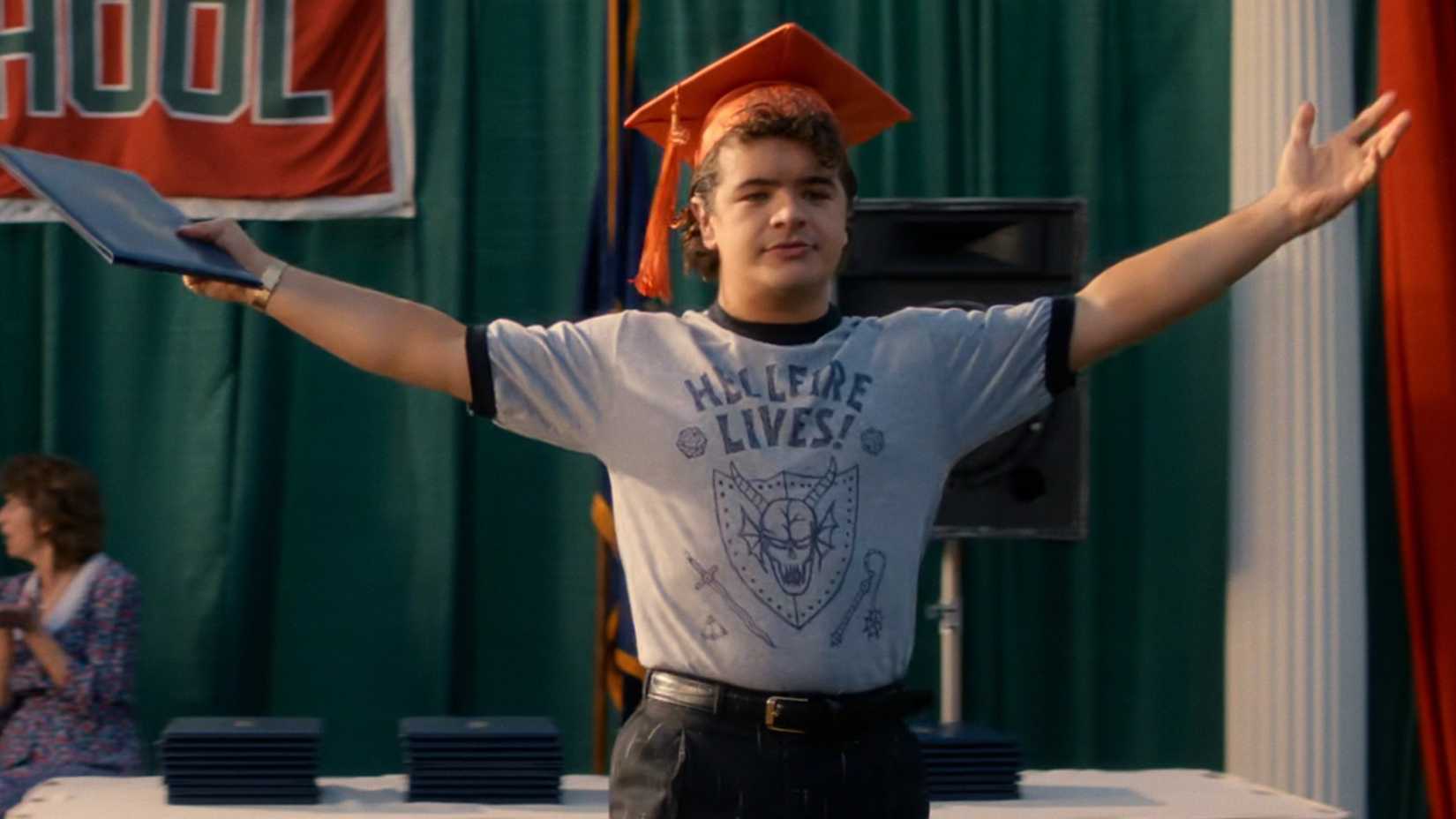 Gaten Matarazzo as Dustin Henderson shows his "Hellfire lives" shirt at the graduation ceremony.
