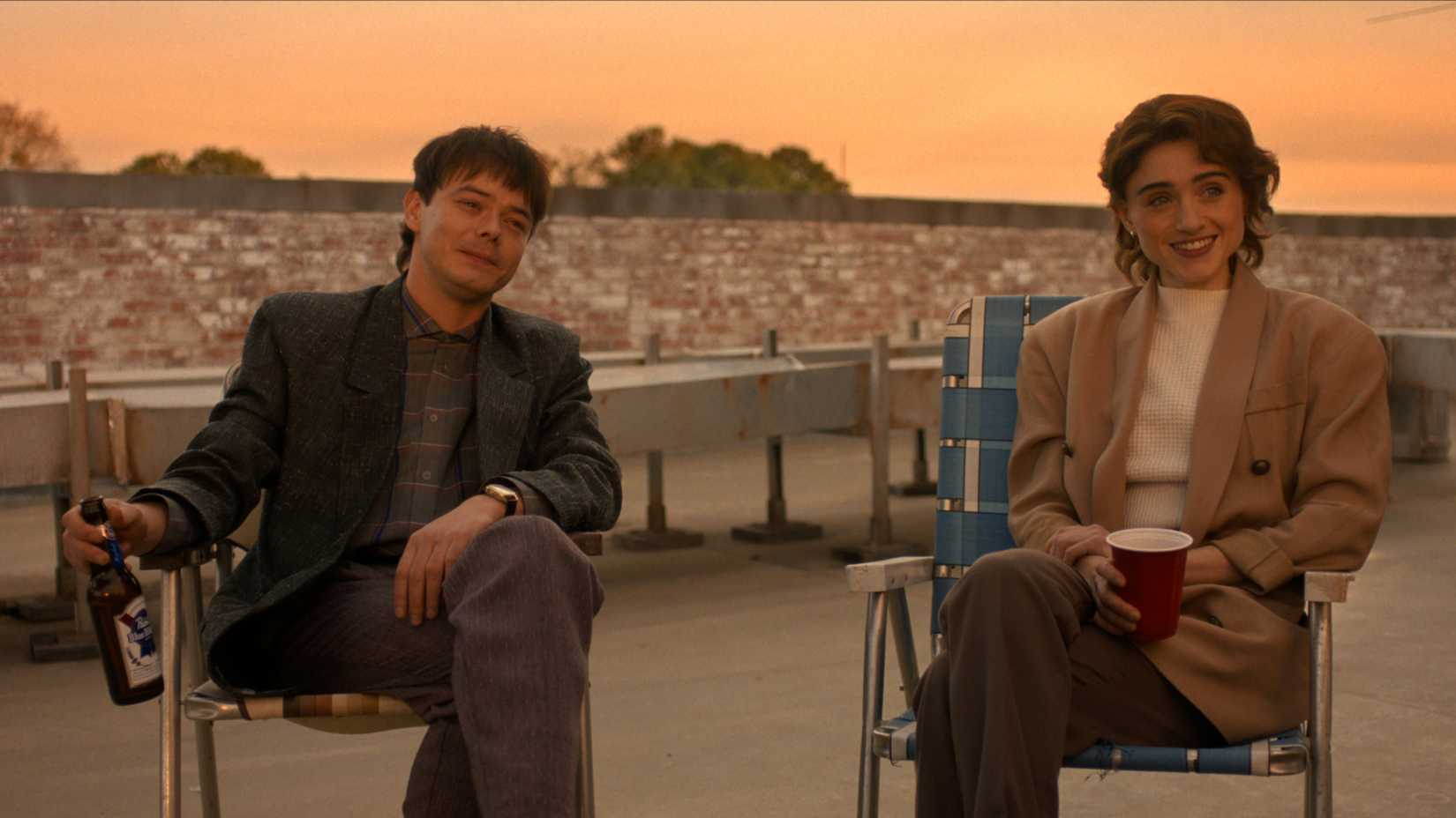 Jonathan and Nancy sitting in lawn chairs drinking and smiling on the roof of the Squawk in Stanger Things season 5 episode 8
