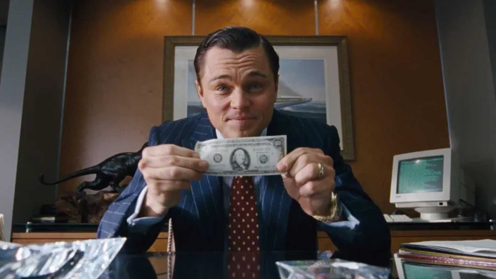 Leonardo DiCaprio como Jordan Belfort segurando uma nota de dólar para a câmera em The Wolf of Wall Street (2013) de Martin Scorsese.