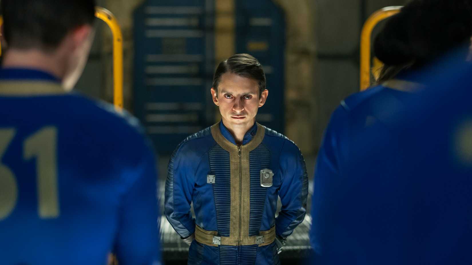 Norm (Moisés Arias) olhando para frente com intensidade enquanto dois habitantes do cofre estão à sua frente em Fallout