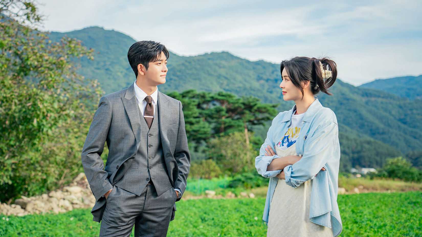 A man in a suit and a woman in casual wear stand in front of a field in the Netflix K-drama The Potato Lab.