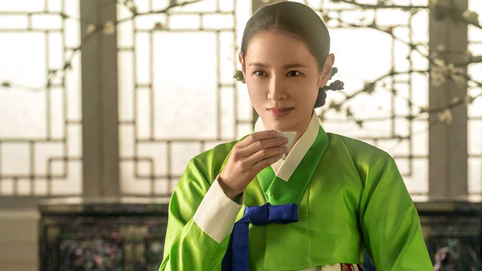 A woman in traditional robes drinks tea while smirking in the historical K-drama The Scandal.