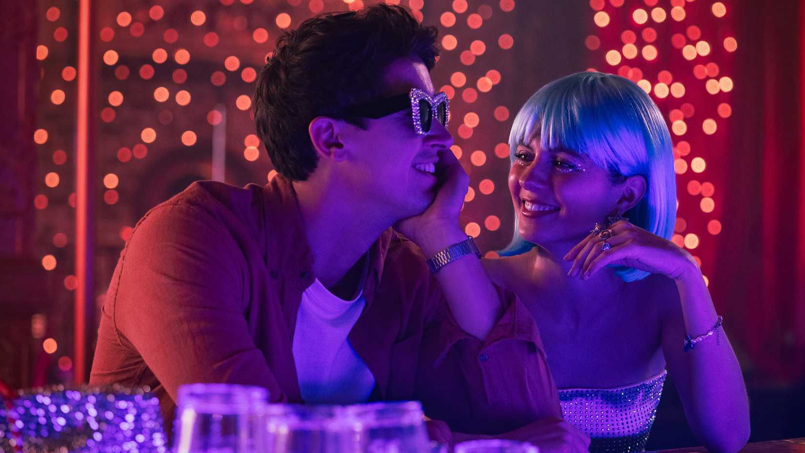 Tom Bryce as Alex Nilsen and Emily Bader as Poppy Wright sit at a bar wearing dressed-up glasses and a wig, among the people they meet on vacation.