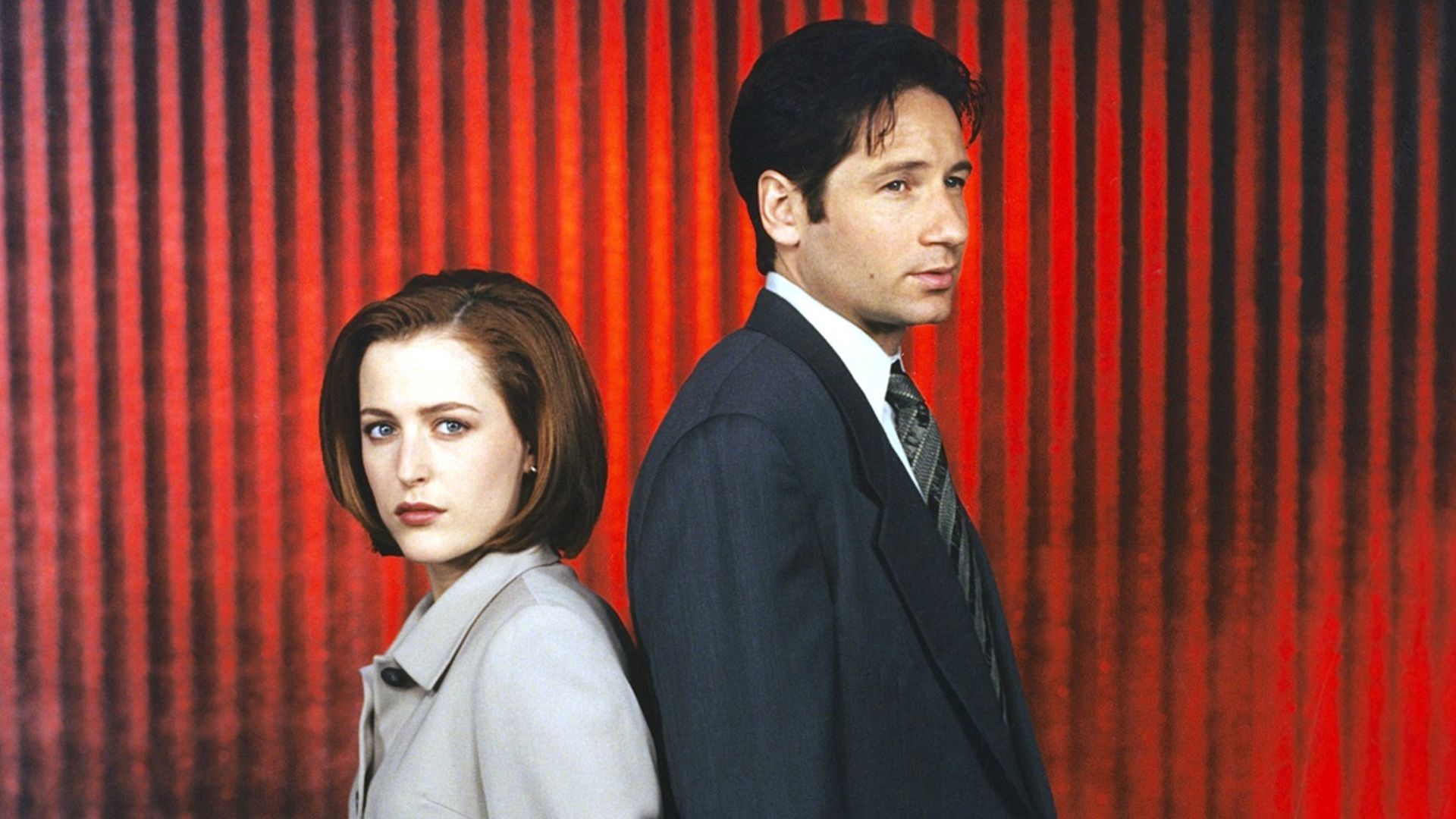 Gillian Anderson and David Duchovny standing together as Mulder and Scully standing together against a red background in a promo photo
