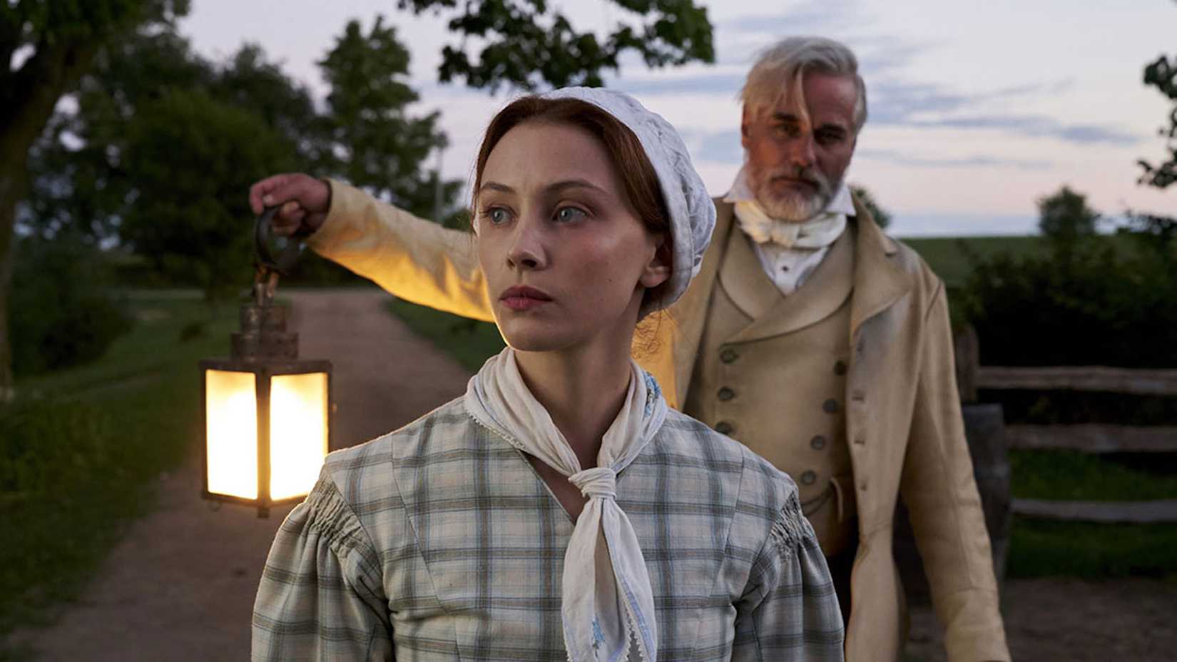 Um homem segurando uma lanterna e uma mulher em trajes coloniais de época em Alias Grace da Netflix.