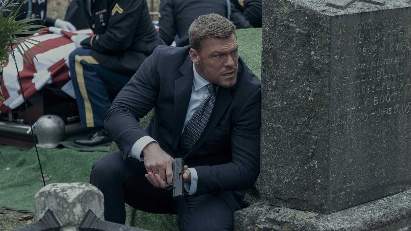 Reacher agachado atrás de uma lápide com uma arma na 2ª temporada de Reacher