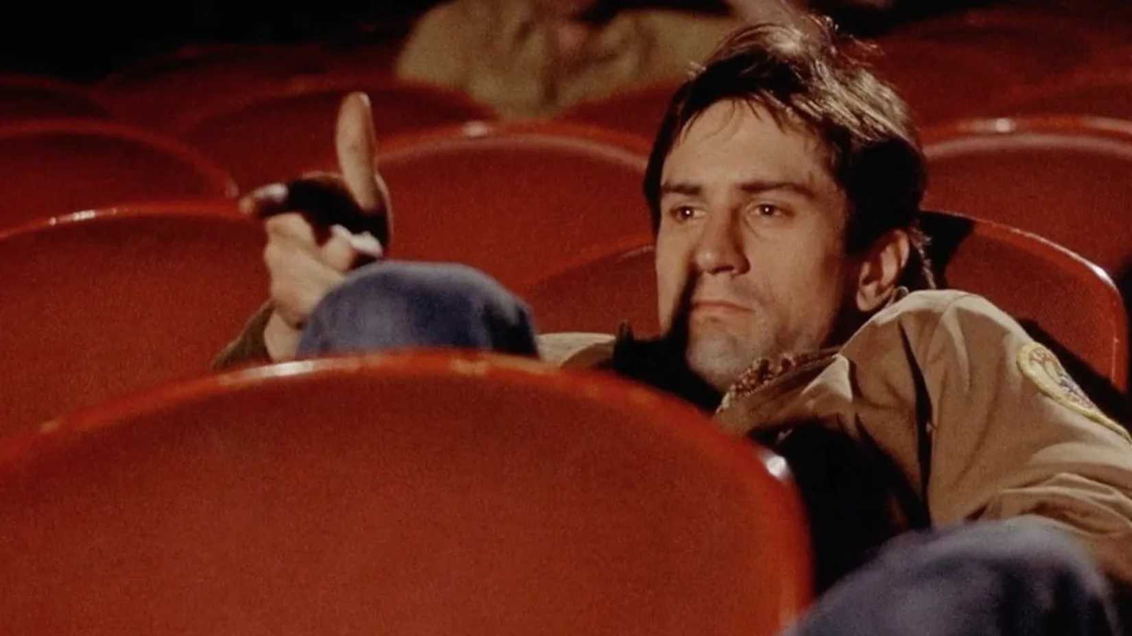 Robert De Niro como Travis Bickle em Taxi Driver fingindo filmar em um cinema.